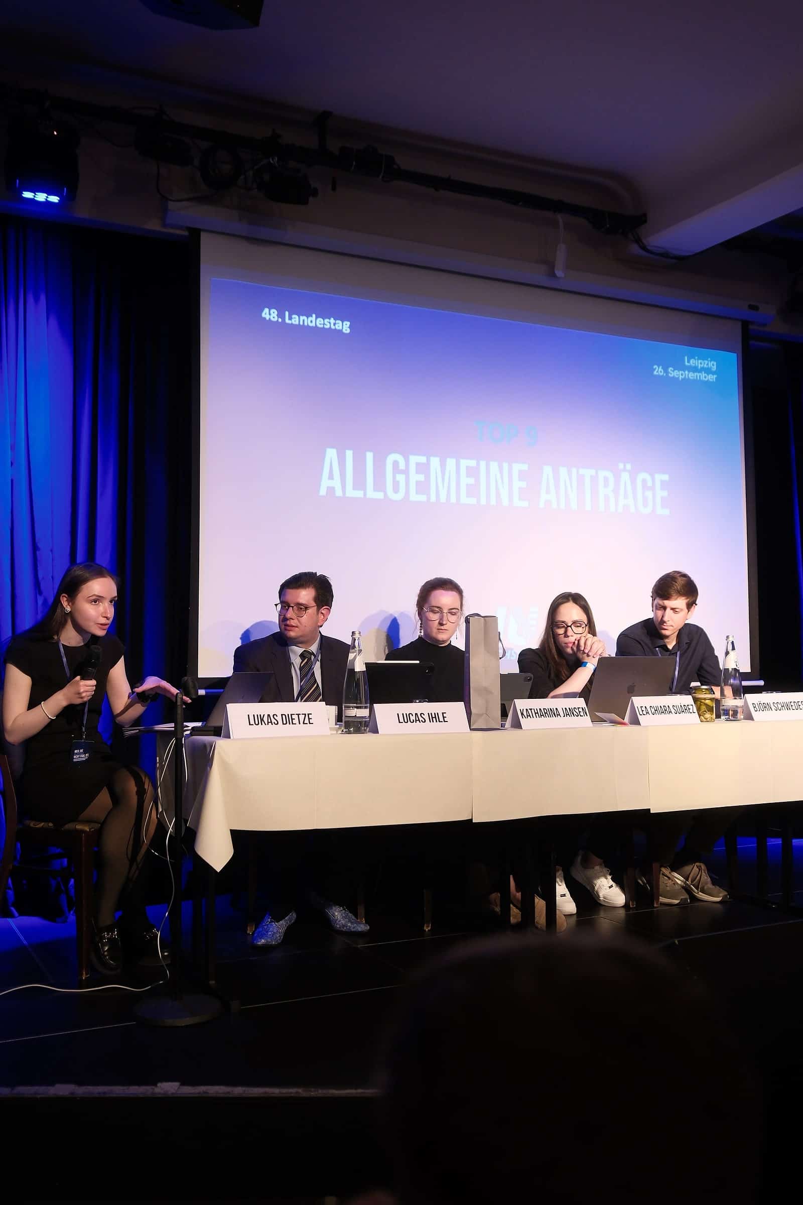 Eine Rednerin spricht neben dem Podium, auf der Leinwand der Punkt Allgemeine Anträge