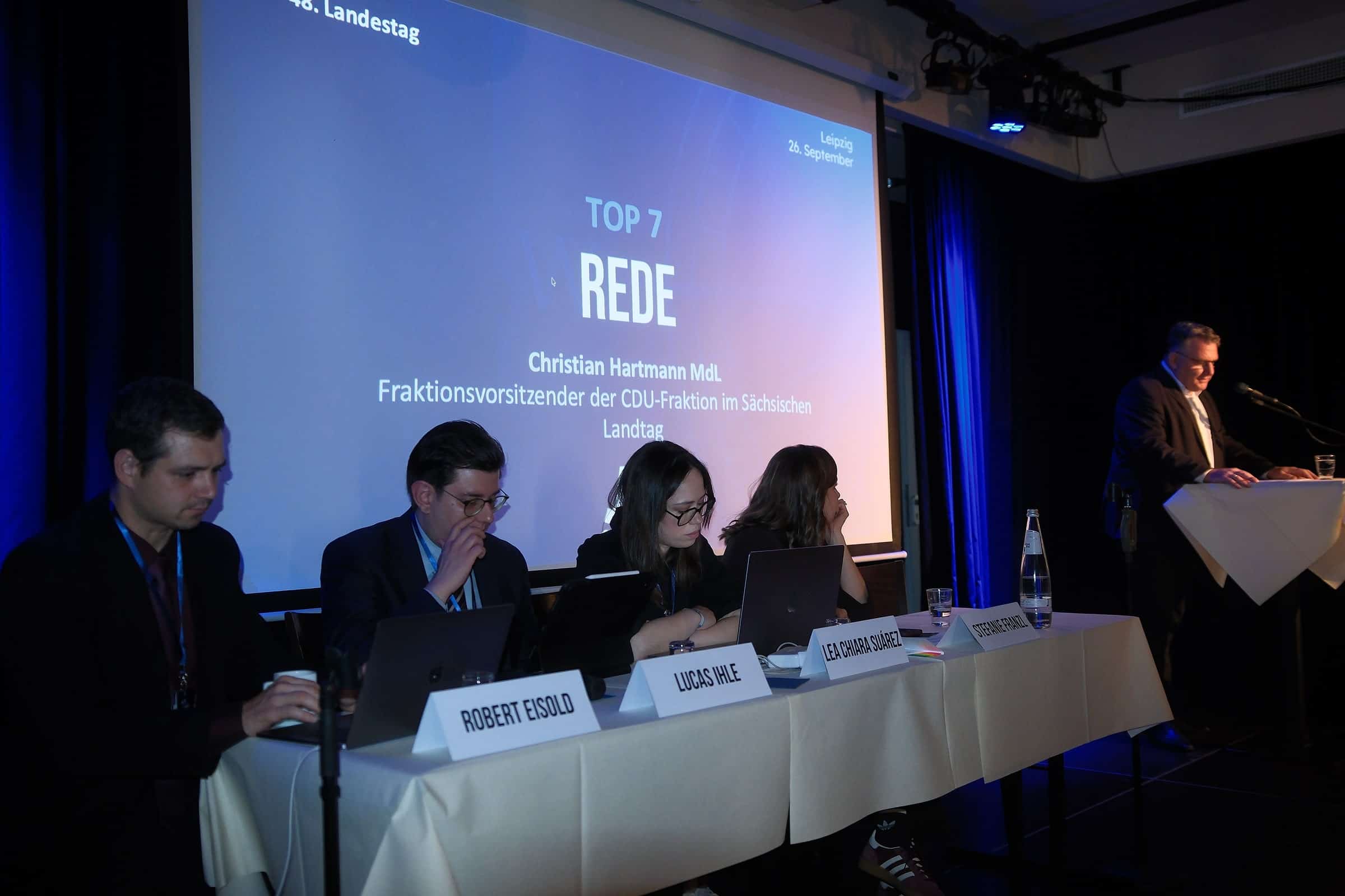 Rede am Pult, daneben das Podium mit aufgeklappten Laptops