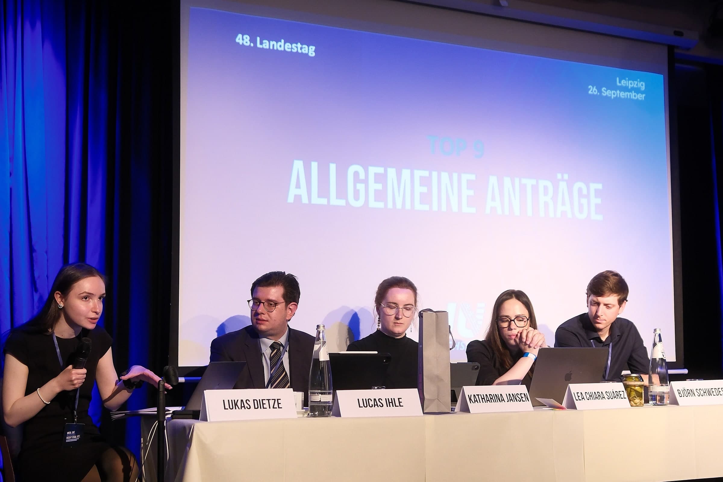 Das Tagungspräsidium auf der Bühne, auf der Leinwand der Tagesordnungspunkt Allgemeine Anträge